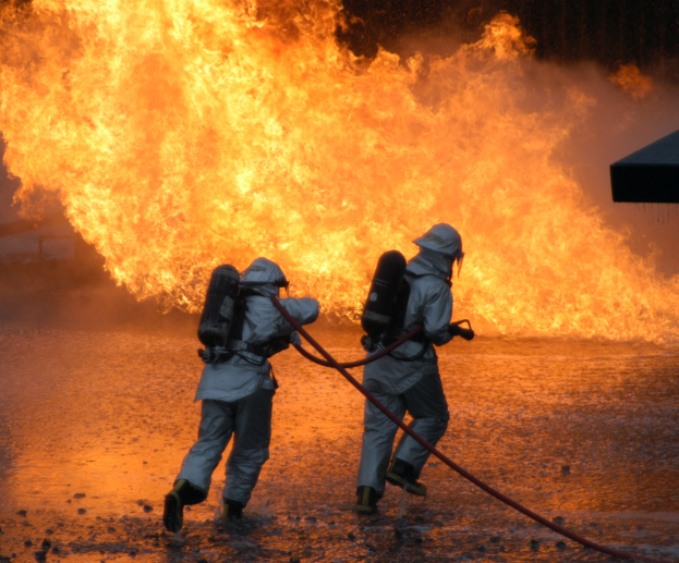 Zwei Feuerwehrleute in Schutzausrüstung verwenden einen Schlauch, um ein Feuer im Hintergrund zu löschen, wobei ein Objekt auf der rechten Seite sichtbar ist.