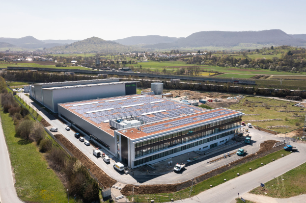 Eine Luftaufnahme eines großen Fabrikgebäudes mit Solarpanelen auf dem Dach, umgeben von Fahrzeugen, Bäumen und anderen Gegenständen, mit Hügeln und einem klaren blauen Himmel im Hintergrund, vermerkt als erstes solarbetriebenes Fabrikgebäude in Deutschland.