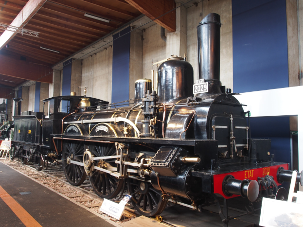 Eine große, alte Dampflokomotive in einem Museums Setting umgeben von Eisenbahnschienen, Informationsplakaten, Ständen und anderen Ausstellungsstücken, mit einer Wand und Deckenbeleuchtung im Hintergrund.