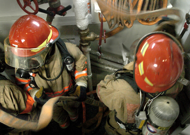 Feuerwehrleute in Helmen und Schutzausrüstung an einem Feuerhydranten mit Rohren, Ventilen und anderen Gegenständen im Hintergrund und Text am unteren Bildrand.