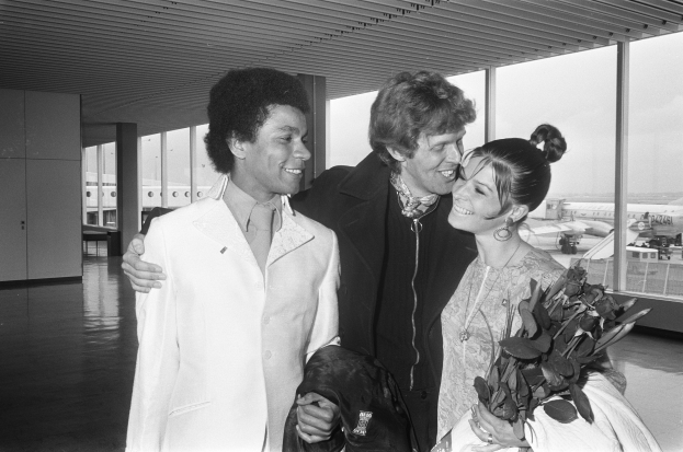 Michael Jackson und seine Frau umarmen und lächeln sich am Flughafen an, Michael trägt einen weißen Anzug und hält eine schwarze Jacke, während seine Frau einen Blumenstrauß hält, Flugzeuge und Gebäude sind im Hintergrund unter einem klaren Himmel zu sehen.