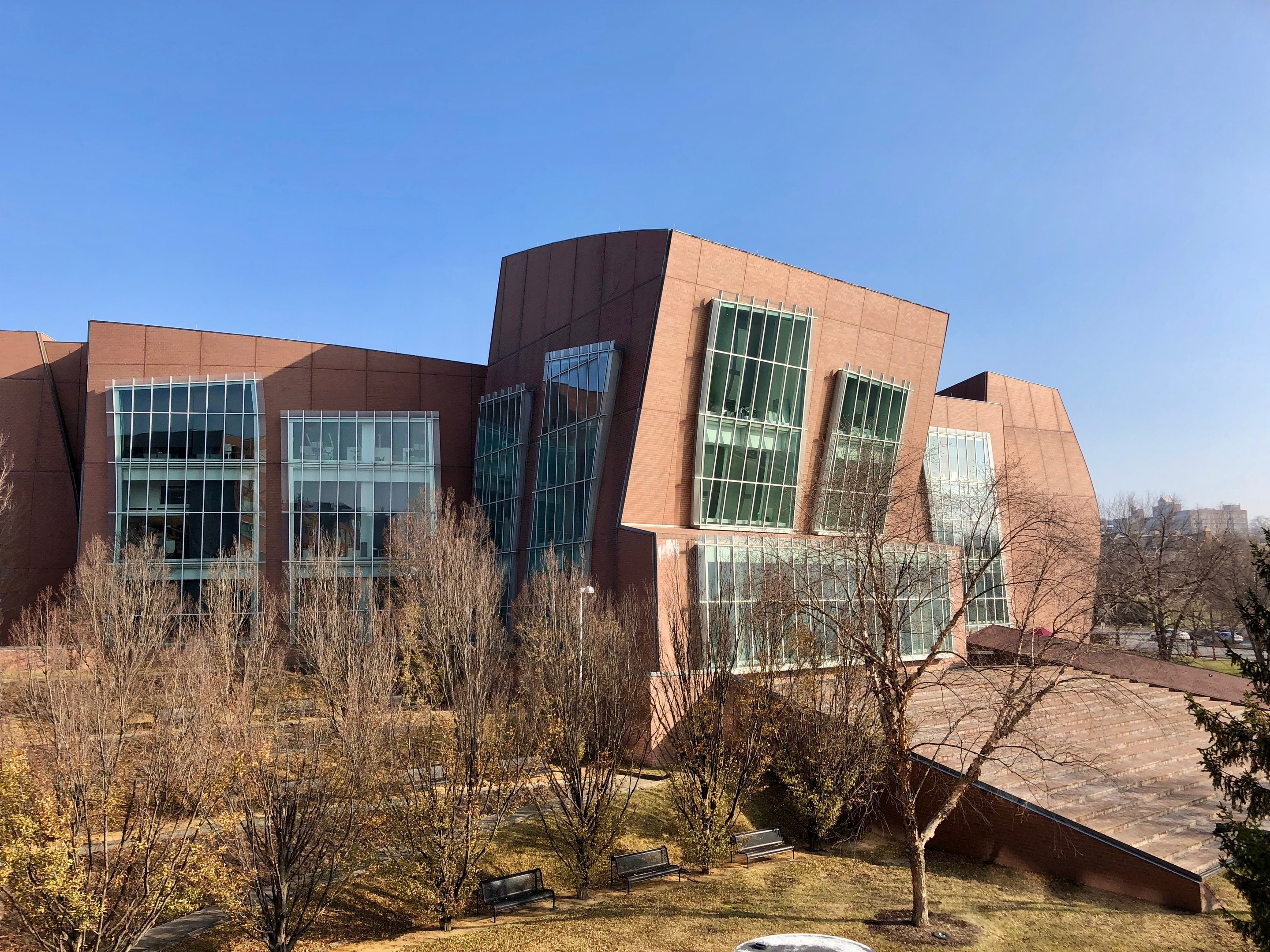 Großes mehrstöckiges Gebäude mit zahlreichen Fenstern, umgeben von Bäumen, Bänken, Gras und einem Laternenmast, vor einem klaren blauen Himmel mit anderen Gebäuden im Hintergrund.