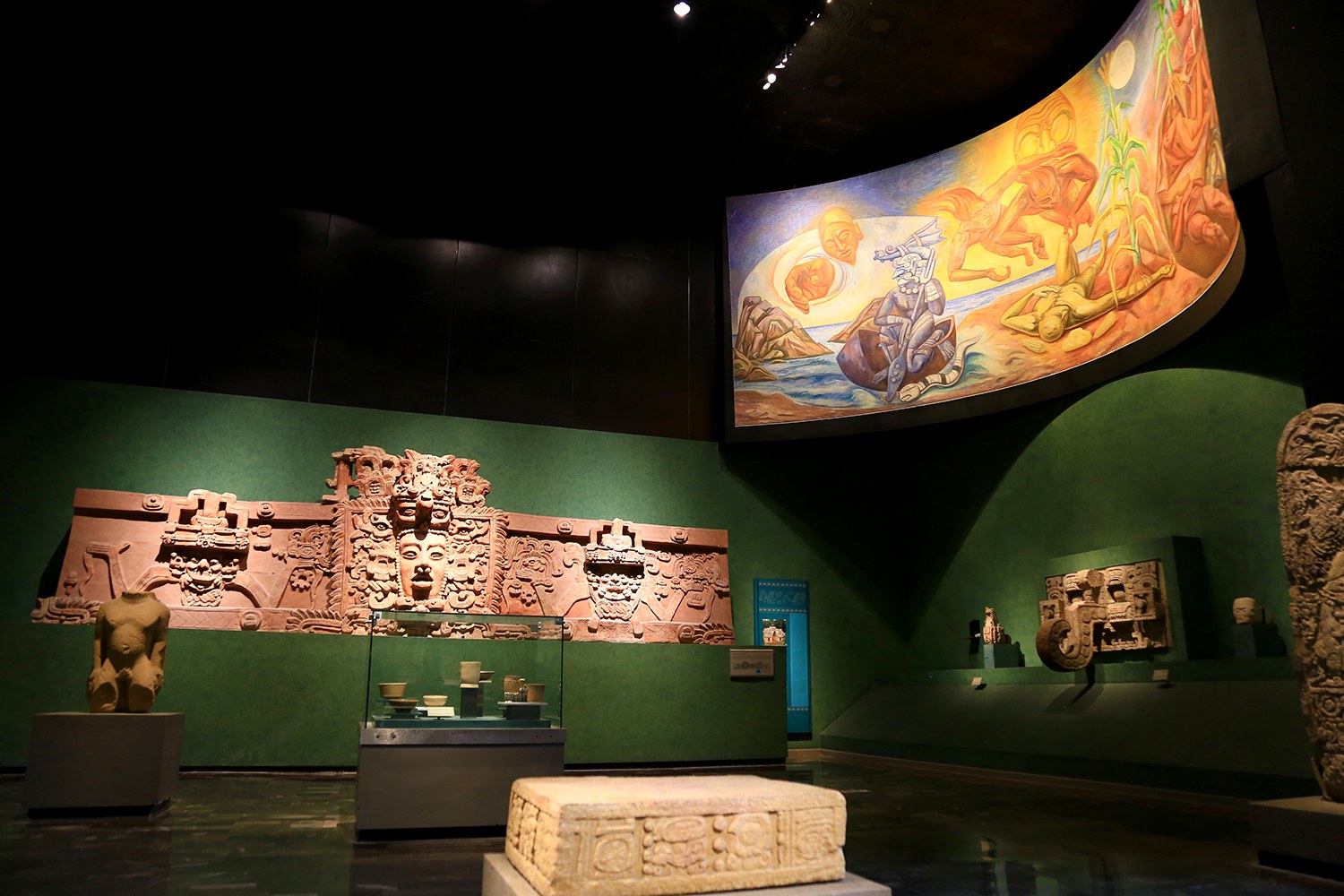 Ein Raum voller verschiedener Artefakte, darunter Skulpturen auf dem Boden, Gegenstände auf Tischen, eine Texttafel, ein Wandgemälde und Deckenleuchten im Mexiko-Stadt-Museum der Anthropologie.