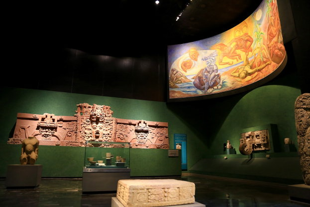 Ein Raum voller verschiedener Artefakte, darunter Skulpturen auf dem Boden, Gegenstände auf Tischen, eine Texttafel, ein Wandgemälde und Deckenleuchten im Mexiko-Stadt-Museum der Anthropologie.