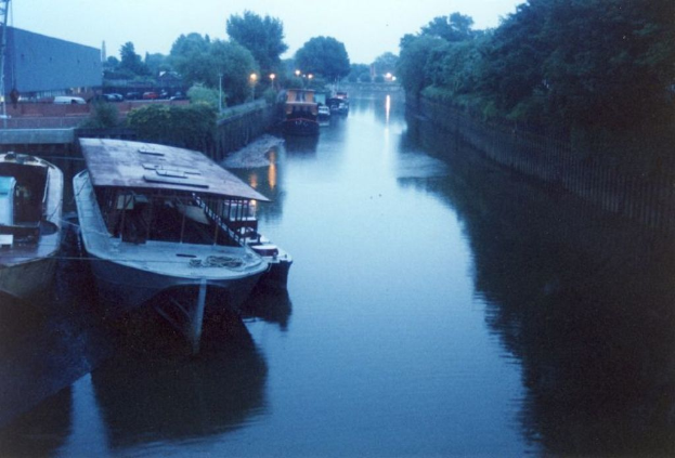 Boote schwimmen in der Nähe des Ufers mit parkenden Fahrzeugen in einer nahen Parkfläche, umgeben von Bäumen und einem Zaun.