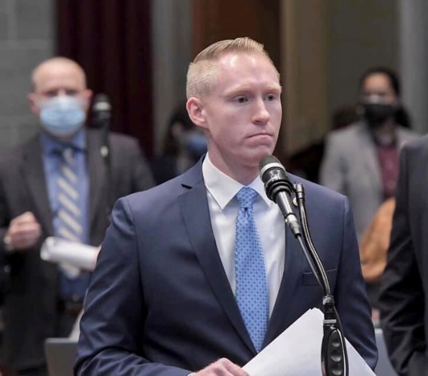 Ein Mann in Anzug und Krawatte spricht an einem Rednerpult mit Papieren in der Hand, während andere in der Nähe stehen.