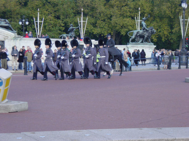 Eine Gruppe von Soldaten in grauen Uniformen marschiert mit Gewehren, begleitet von einem Mann auf einem Pferd, während Zuschauer von der Seite zusehen, mit Statuen und Bäumen im Hintergrund.