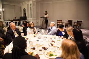 Gruppe von Menschen um einen Esstisch mit Essen und Besteck in einem Raum mit einer Frau an einem Podium, einer weiteren Person in der Nähe eines Geländers und Tischen mit Flaschen und Beleuchtung.