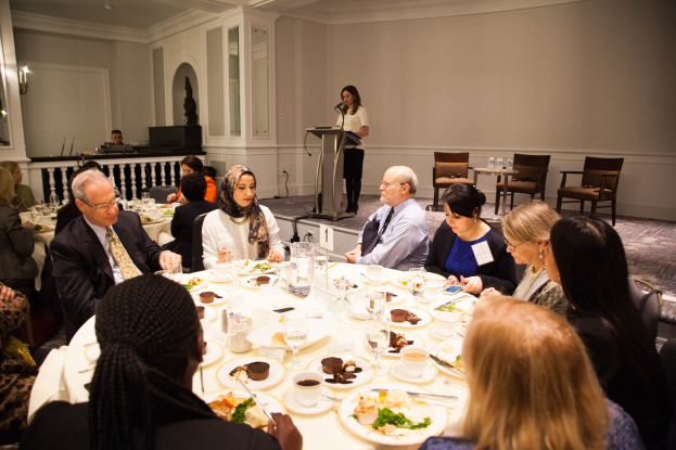 Gruppe von Menschen um einen Esstisch mit Essen und Besteck in einem Raum mit einer Frau an einem Podium, einer weiteren Person in der Nähe eines Geländers und Tischen mit Flaschen und Beleuchtung.