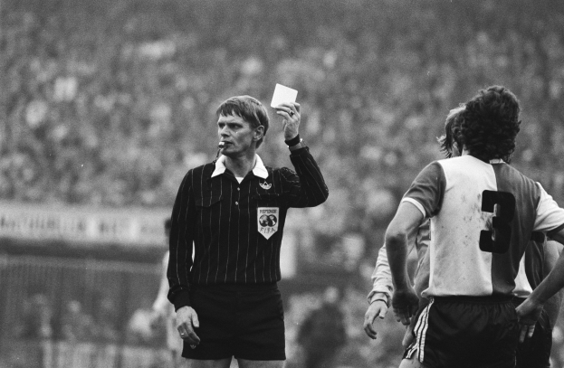 Schwarzes und weißes Bild eines Schiedsrichters, der eine rote Karte in der Hand hält und vor einer Menge von Menschen steht, die Fußballuniformen tragen, wobei ein Zaun im Hintergrund zu sehen ist.