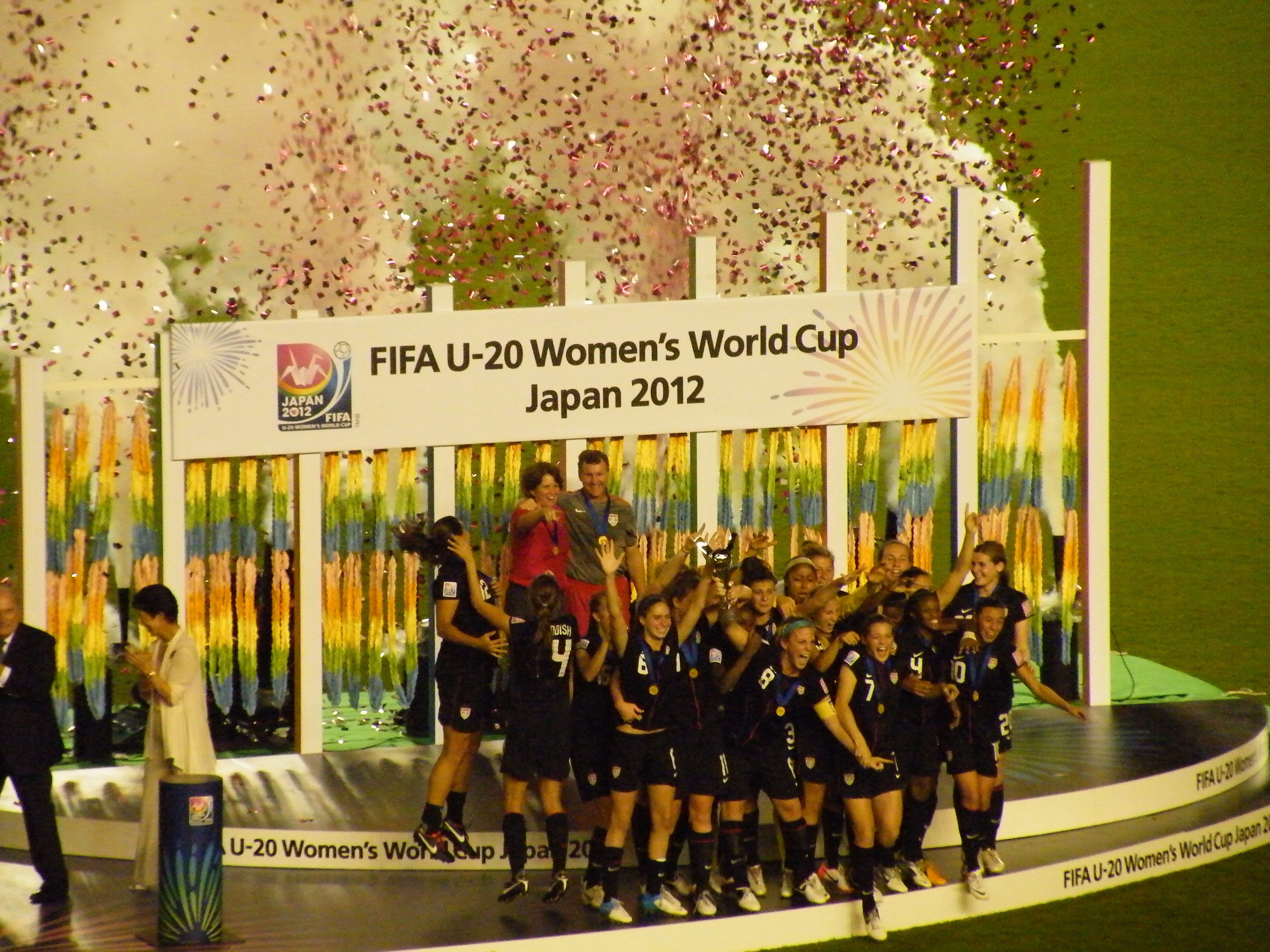 Schwarze Frauenfußballspielerinnen feiern auf einer Bühne mit Publikum und dekorativem Hintergrund.