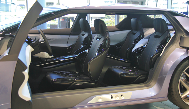 Das Innenleben eines Autos mit öffnender Tür, das luxuriöse Ledersitze, Lenkrad und Räder zeigt, mit Gebäuden und Bäumen im Hintergrund.