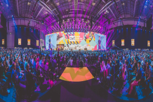 Eine große Menschenmenge sitzt auf Stühlen vor einer hell erleuchteten Bühne mit mehreren Personen, die wahrscheinlich an einem Online-Marketing-Rockstars-Event teilnehmen, umgeben von Bildschirmen.