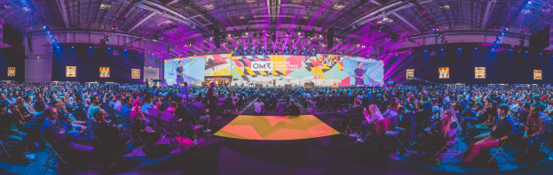 Eine große Menschenmenge sitzt auf Stühlen vor einer hell erleuchteten Bühne mit mehreren Personen, die wahrscheinlich an einem Online-Marketing-Rockstars-Event teilnehmen, umgeben von Bildschirmen.