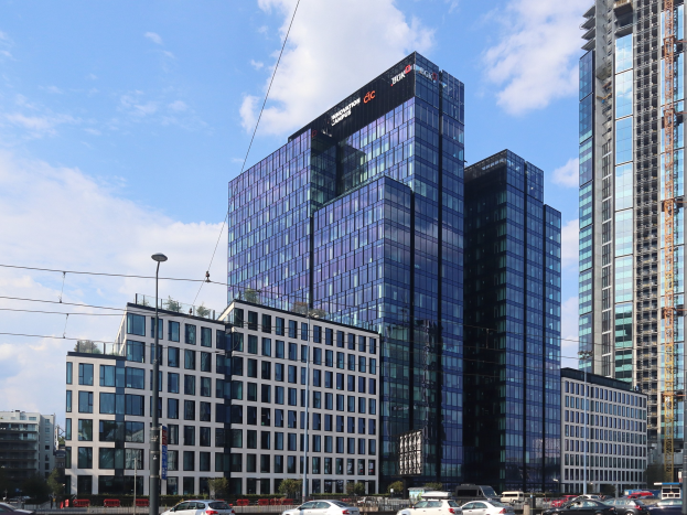 Außenansicht des Deutschen Bank-Hauptsitzes in Frankfurt, Deutschland, mit einem großen Gebäude mit vielen Fenstern, umgeben von städtischer Infrastruktur und Grünflächen unter einem bewölkten Himmel.