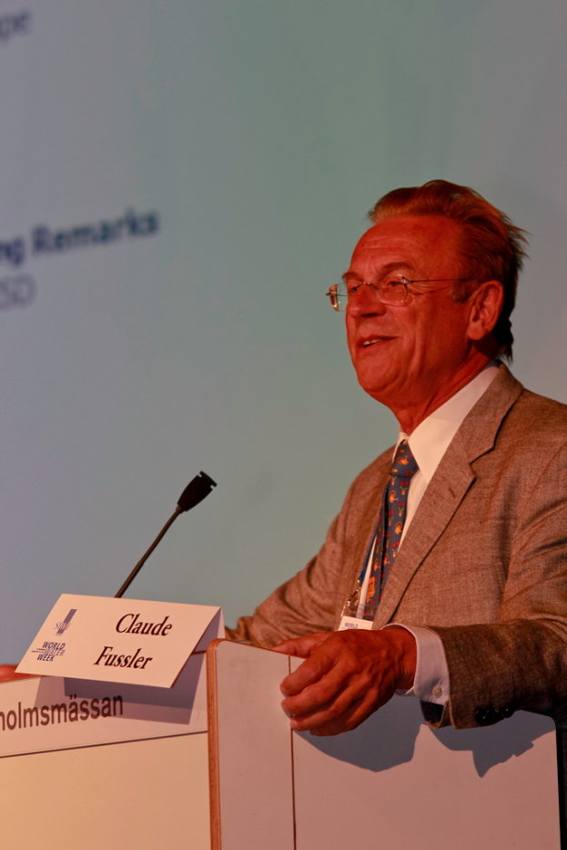 Ein Mann in einem weißen Hemd und einem grauen Blazer steht lächelnd vor einem Podium mit Mikrofon und Namensschildern, mit einem Projektionsschirm oder einer Banner im Hintergrund.