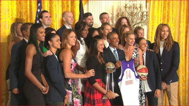 Präsident Obama und First Lady Michelle Obama posieren mit dem Damen-Basketball-Team im Oval Office des Weißen Hauses, halten einen Basketball, eine Trophäe und lächeln mit einer Flagge, Vorhängen und einer Kerzenleuchter im Hintergrund.