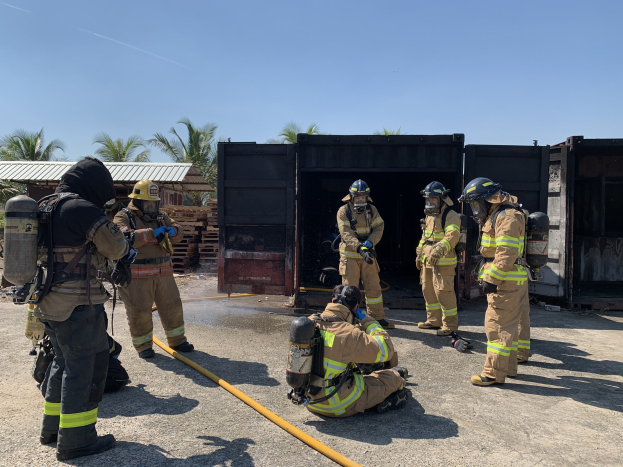 Feuerwehrleute in Helmen und Ausrüstung stehen vor einem Gebäude und halten Rohre, mit Bäumen, Schuppen und hölzernen Gegenständen im Hintergrund während einer scheinbar Feuerlöschtrainingssitzung.