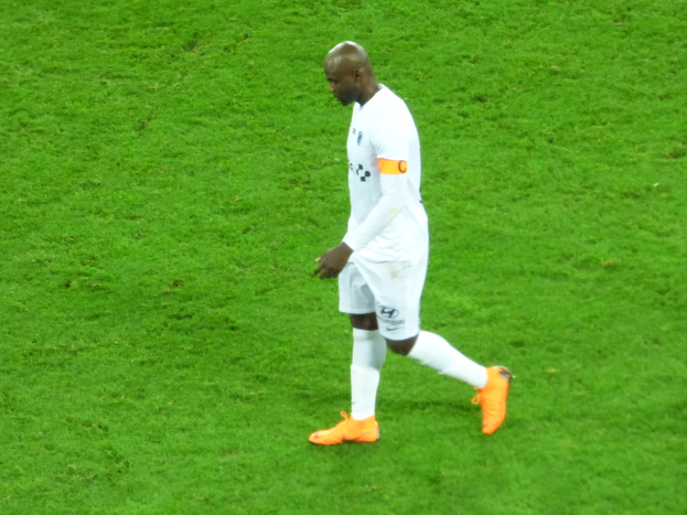 Ein Fussballspieler in einem weissen T-Shirt, weissen Shorts und orangen Schuhen läuft über ein saftiges grünes Feld.
