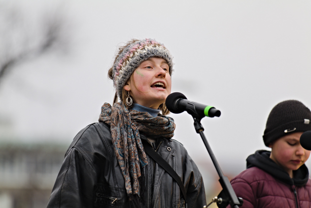 Eine Frau in schwarzer Jacke, Schal und Mütze singt in ein Mikrofon auf der Bühne, neben ihr eine Person in einer maronenfarbenen Jacke und schwarzer Mütze, während ein unscharfer Hintergrund die Menge bildet.