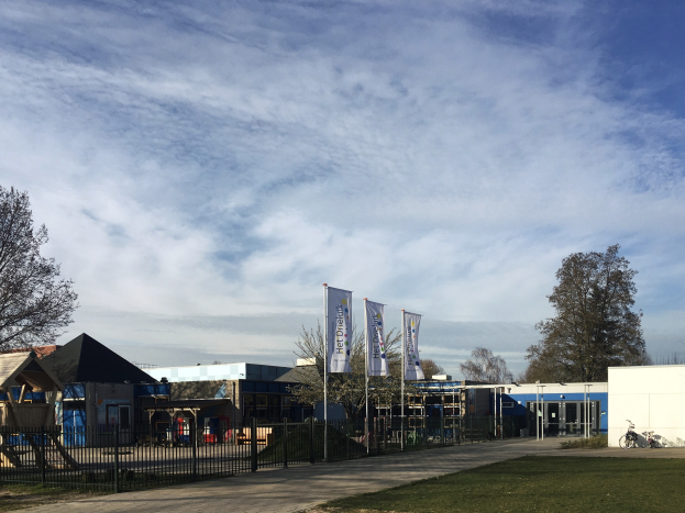 Großes Gebäude mit Metallzaun, Rasenfläche, Weg, Bäumen, Bannern, Fahrrad, nahen Häusern und bewölktem Himmel, als neuer Spielbereich für Kinder an der Schule identifiziert.