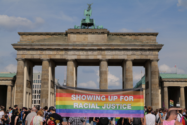 Eine Gruppe von Menschen hält eine 'Racial Justice'-Tafel vor dem Brandenburger Tor in Berlin, mit den Säulen und der Statue des Tors im Hintergrund, vor Gebäuden und einem bewölkten Himmel.