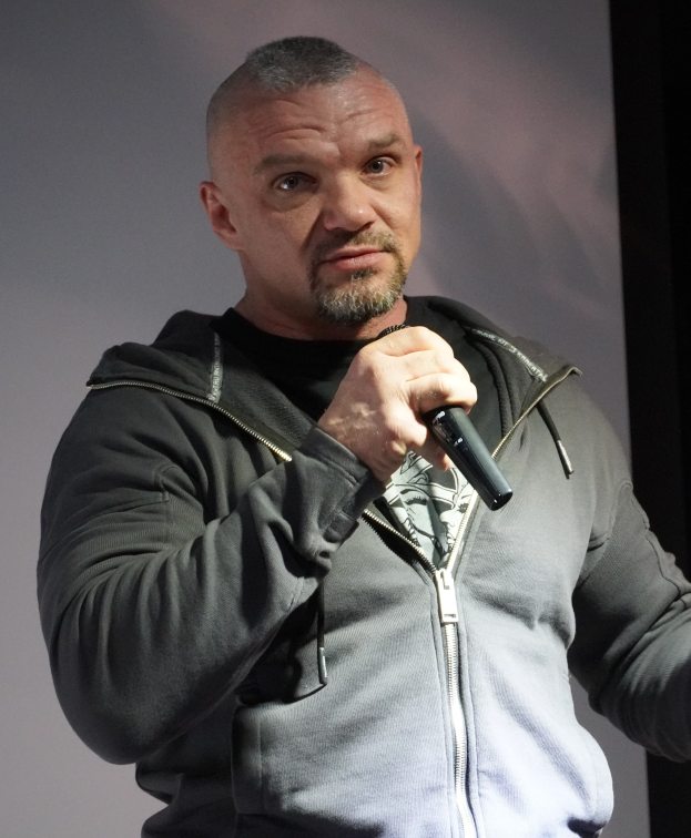 Ein Mann in einer schwarzen Kapuzenjacke und einem schwarzen Anzug mit einem weißen Hemd, der ein Mikrofon hält, steht selbstbewusst vor einer Wand und sieht aus wie ein Comedian.