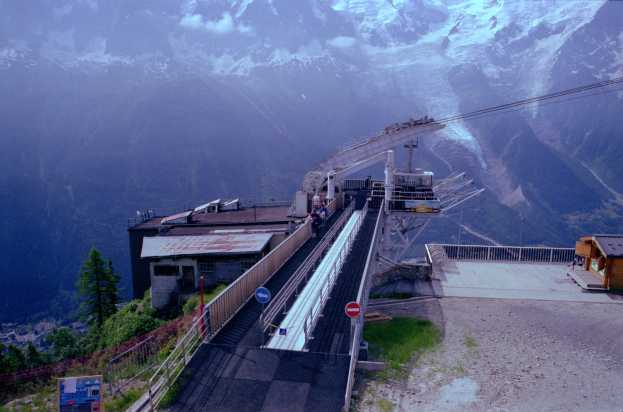 Seilbahn fährt einen schneebedeckten Berg hinauf, im Hintergrund sind weitere schneebedeckte Gipfel zu sehen, unten verläuft eine Straße mit Schildern, Geländern, Vegetation und Häusern, sowie einigen Menschen.