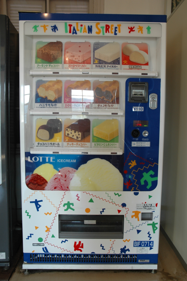 Ein Snackautomat mit einer Vielzahl von Snacks, einem Münzschlitz oben und einem Spender unten, der an einer Wand mit einem Fenster im Hintergrund steht.
