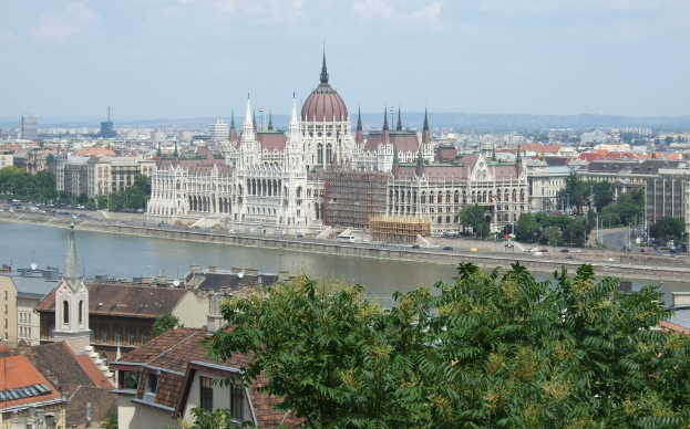 Ein Fluss durchquert die Szene mit Gebäuden und Bäumen auf der linken Flussseite, einer Straße mit fahrenden Fahrzeugen und Masten auf der rechten Seite und einem klaren Himmel mit einem Berg im Hintergrund.