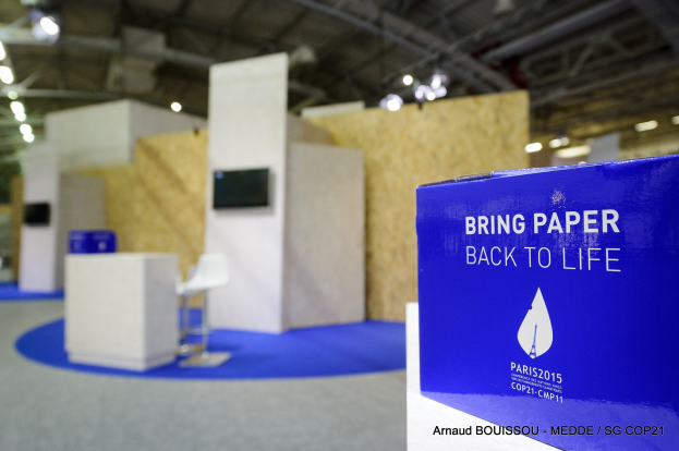 Ein Messestand mit einem blauen und weißen 'Bring Paper Back to Life'-Schild auf der rechten Seite, der blasse Stühle, Tische, einen wandmontierten Fernseher und Deckenleuchten im Hintergrund zeigt.