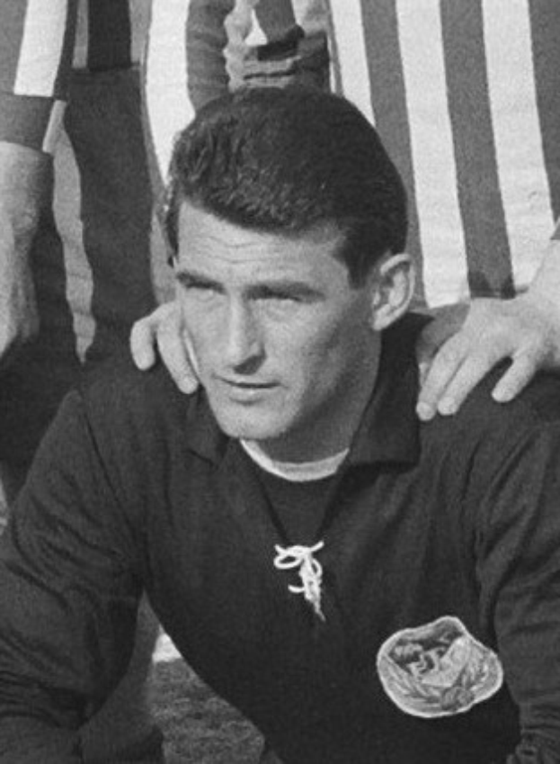 Ein Schwarz-Weiß-Foto eines Mannes, der auf einem Rasenfußballfeld sitzt und ein T-Shirt sowie eine Kette trägt, während zwei Personen hinter ihm stehen.