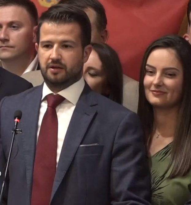 Mann im Anzug und Krawatte spricht vor einem Publikum mit Mikrofon, mit einem Banner im Hintergrund.