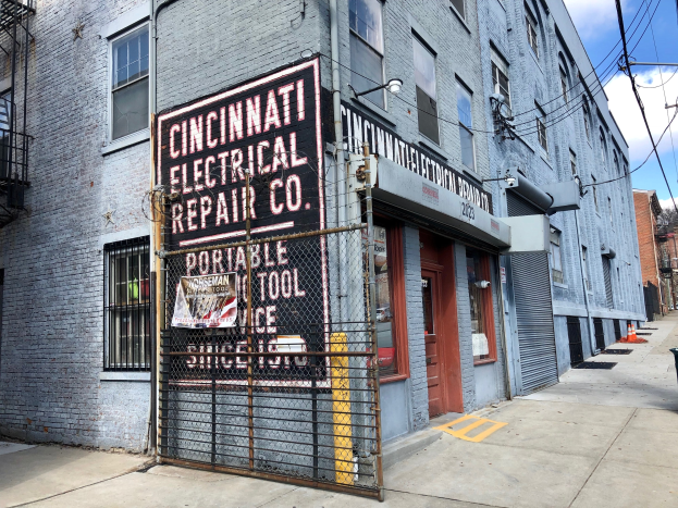 Außenansicht des Cincinnati Electrical Repair Co.-Gebäudes mit einem Schild, einem Metallzaun, einem Müllcontainer, Strommasten, Drähten, Bäumen und einem bewölkten Himmel.