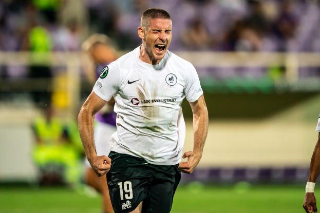 Ein Mann in weißem Hemd und schwarzen Shorts rennt auf einem Fußballfeld, feiert ein Tor, umgeben von einer Gruppe von Menschen, mit unscharfem Hintergrund.