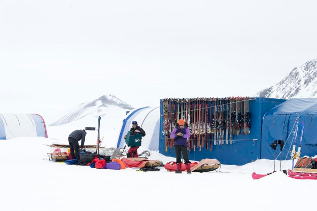 Drei Personen stehen auf einer schneebedeckten Landschaft mit verstreuten Taschen, Zelten mit Skiern dahinter und schneebedeckten Höhen im Hintergrund unter einem klaren Himmel.