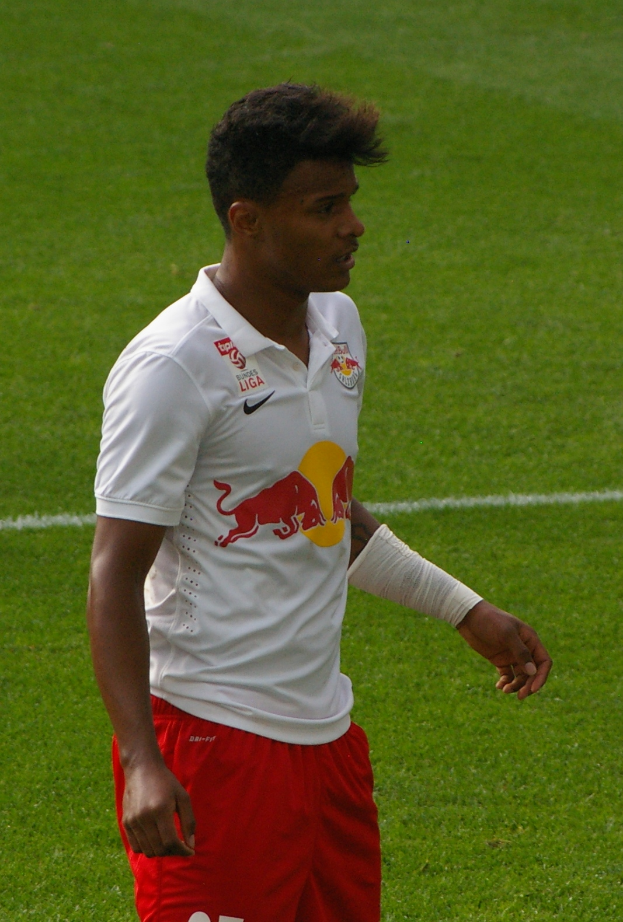 Ein Mann in einem weißen Hemd und roten Shorts steht auf einem Fußballfeld und schaut rechts mit entschlossener Miene, er scheint ein Flügelstürmer zu sein.