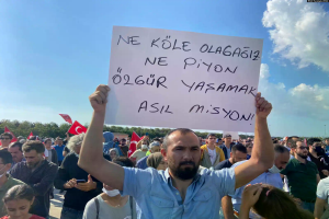 Ein Mann hält ein Schild mit der Aufschrift "Aserbaidschanische Protestler demonstrieren in Ankara für den Rücktritt von Präsident Ilham Aliyev" vor einer großen Menge mit Fahnen und Handys, unter einem blauen und weißen Himmel mit Bäumen im Hintergrund.