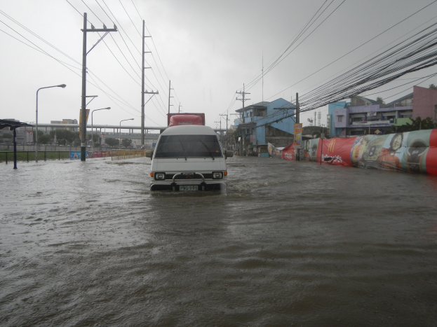 Ein Van fährt durch eine überflutete Stadtstraße mit Strommasten, Laternen und Gebäuden auf beiden Seiten, eine Brücke im Hintergrund und der Himmel darüber.