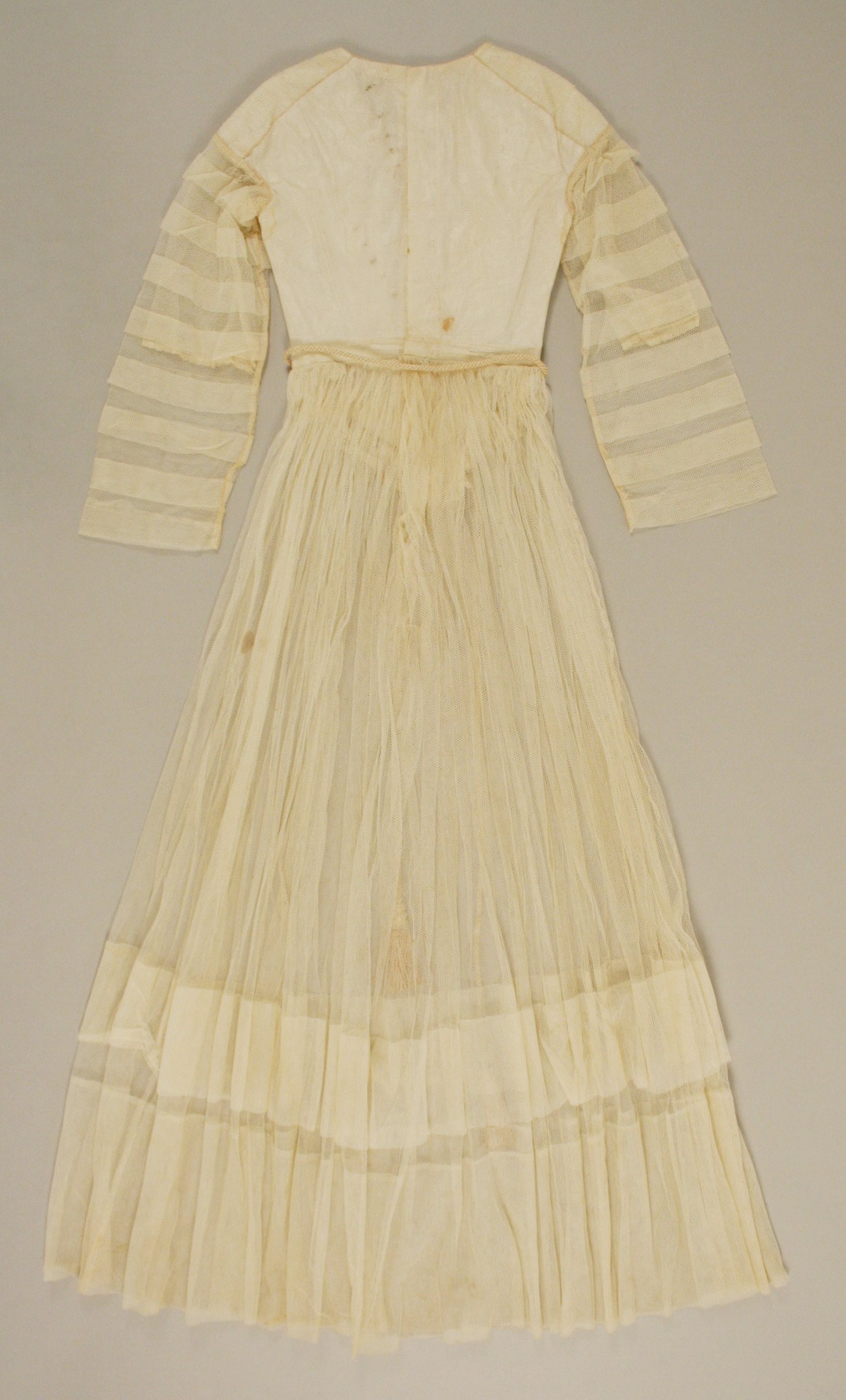 Ein weißes, langärmeliges Kleid mit einer gerüschten, bodenlangen Schleppe und hohem Bund auf einem weißen Hintergrund, das aus einem leichten, luftigen Stoff mit einem dezenten Schimmer besteht.