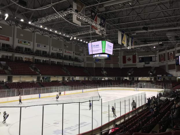 Eishockeyspiel in einer großen Arena mit einem zentralen Netz, Zuschauern auf Stühlen, Deckenleuchten und Trägern sowie Banner an den Wänden, die "St. Louis Blues Arena" beschriften.