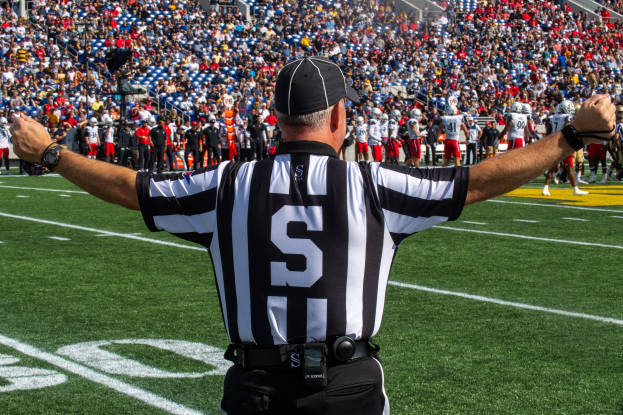 Ein Schiedsrichter steht auf einem Fußballfeld mit ausgestreckten Armen, umgeben von einer Gruppe von Menschen auf dem Boden und einer sitzenden Menge im Hintergrund.