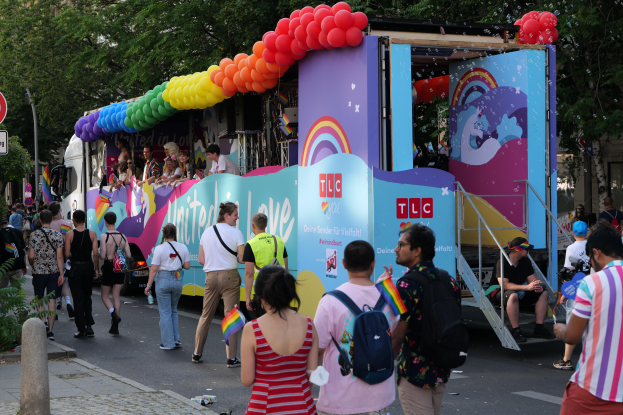 Eine Gruppe von Menschen, die auf einer Straße neben einem Lastwagen mit bunten Luftballons gehen, mit Schildern an Pfosten auf der Straße und Bäumen und Gebäuden im Hintergrund, was auf eine Pride-Parade in Paris hindeutet.