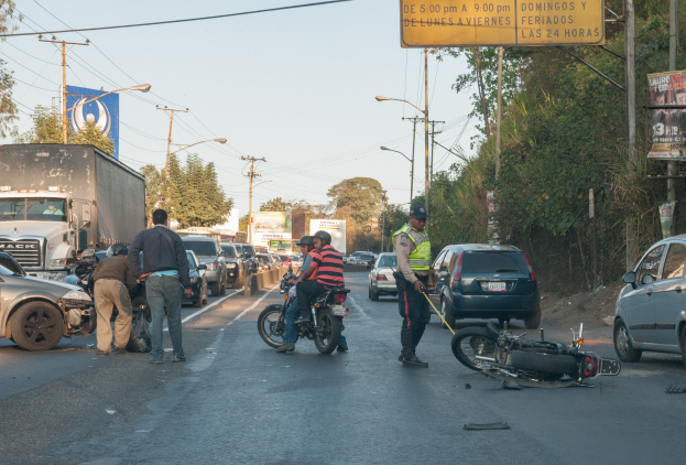 Gruppe von Menschen um ein verunglücktes Motorrad auf der Stra√enseite mit mehreren Fahrzeugen, darunter ein Lastwagen, und Hintergrundelementen wie Bäumen, Masten, Lichter, Schilder und Himmel.