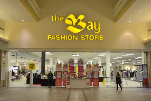Verkaufsfläche eines Bay Fashion-Geschäfts in einem Einkaufszentrum mit vorbeigehenden Kunden, Informationsschilder an den Wänden, tragenden Säulen, Deckenbeleuchtung und zusätzlichen Mall-Elementen im Hintergrund.