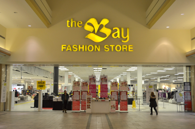 Verkaufsfläche eines Bay Fashion-Geschäfts in einem Einkaufszentrum mit vorbeigehenden Kunden, Informationsschilder an den Wänden, tragenden Säulen, Deckenbeleuchtung und zusätzlichen Mall-Elementen im Hintergrund.