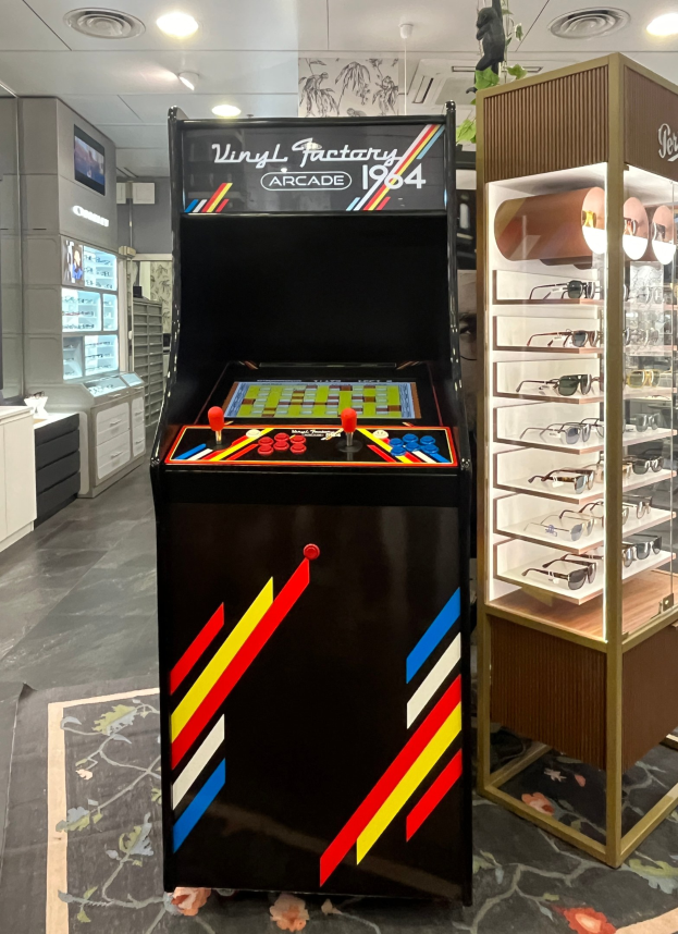 Ein Geschäft mit einem zentralen Vintage-Arcade-Automaten, einem Gestell mit Gläsern auf der rechten Seite und Hintergrund-Elementen wie Schränken, einem Fernseher und anderen Gegenständen, wobei Deckenleuchten für eine retro-mäßige Atmosphäre sorgen.