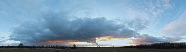 Ein bewölkter Himmel mit einem Fabrikschornstein, der Rauch abgibt, und ein paar Bäumen und Pflanzen auf der rechten Seite.