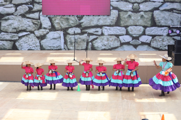 Gruppe junger Mädchen in bunten Kleidern und Hüten, die auf einer Bühne mit Verkehrskegeln, Mikrofonen, Lautsprechern, Scheinwerfern und einem Bildschirm mit Text im Hintergrund tanzen, während das Publikum unten zusieht.