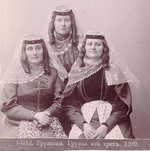 Drei Frauen in traditionellen russischen Hochzeitskleidern mit aufwendiger Stickerei und Perlen stehen vor einer weißen Wand, mit Text unten, der darauf hinweist, dass es ein russisches Hochzeitsfoto ist.